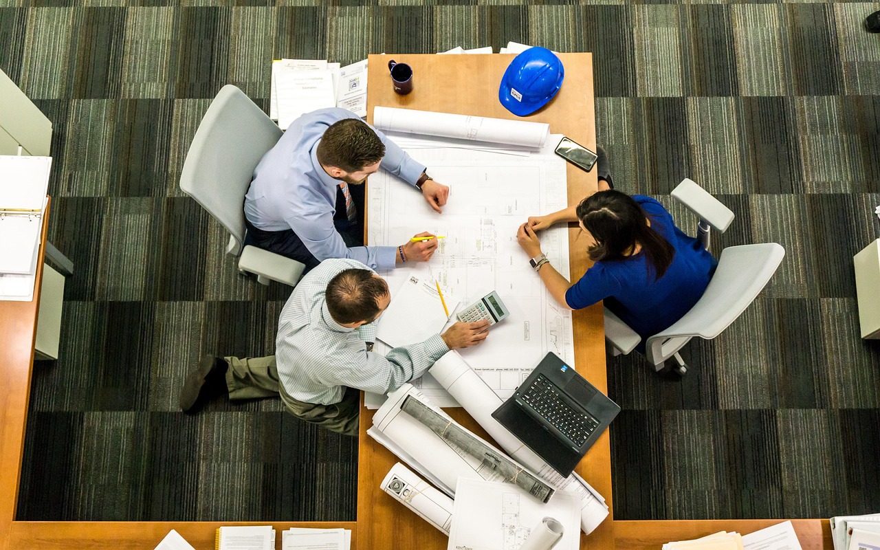 HOA board meeting—three people review building plans with a laptop, calculator, and blue hard hat, symbolizing Red Brick’s support.