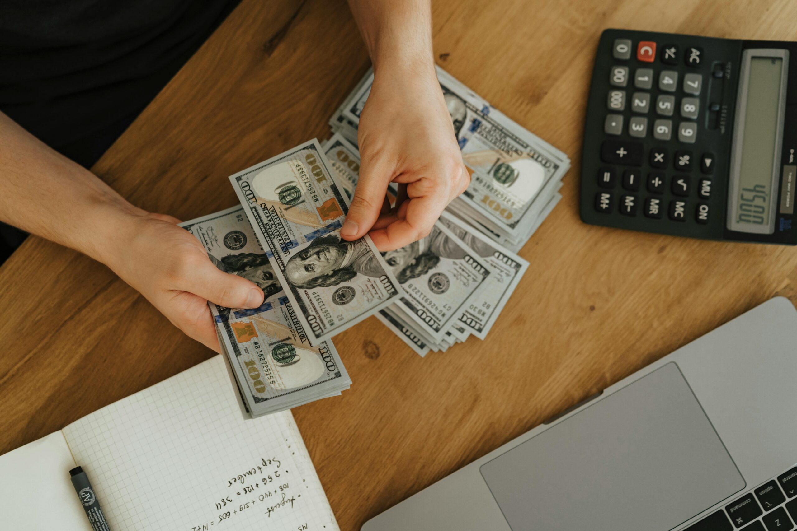 Person counting US dollar bills on a desk with a calculator, notebook, and laptop, symbolizing financial planning and reserve fund management.